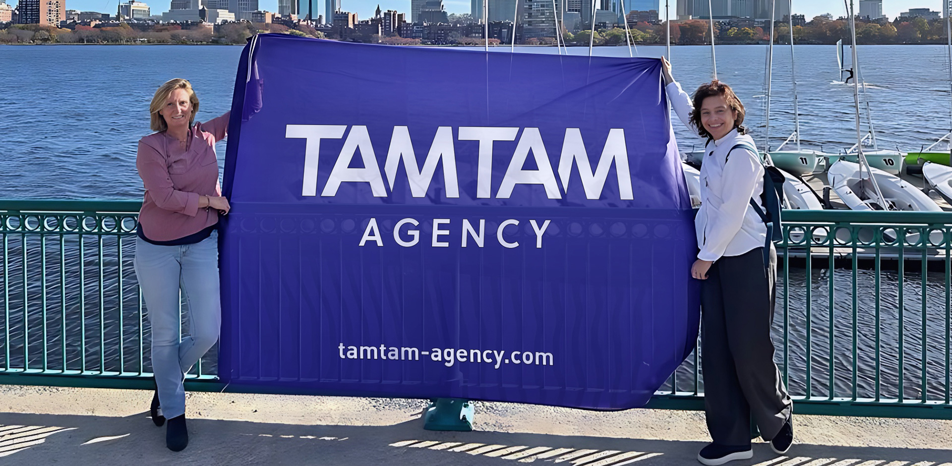 Beatrice and Karine hold the TAM TAM flag in front of the Charles River